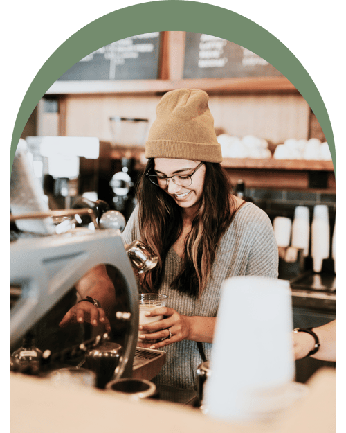 A barista in a cozy café, wearing a yellow beanie, prepares a drink behind a coffee machine, with pastries on display above. A barista in a cozy café, wearing a yellow beanie, prepares a drink behind a coffee machine, with pastries on display above.