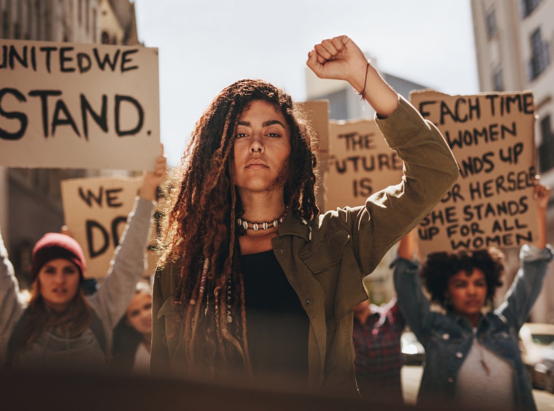 a scene of a protest with a woman in the centre facing toward camera with fist raised in the air